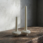Two white candle holders with candles on a rustic wooden surface against a textured wall.