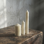Three white candles of different shapes on a wooden surface with a textured wall background